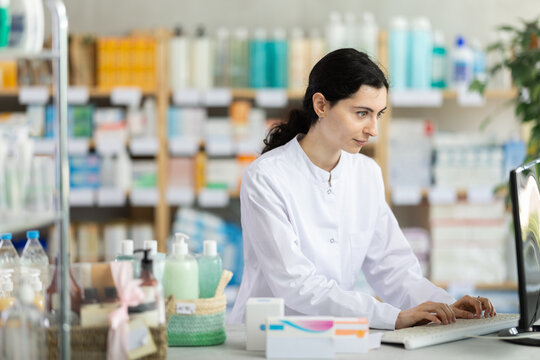 Female pharmacist in white medical coat working at computer in pharmacy