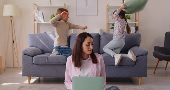 Mother sits with her laptop, trying to focus on her work amidst commotion from her children. Woman holds head in annoyance, trying to find peace as noisy children playfully fight pillows behind her. 