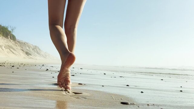 Close up of a woman walking barefoot on sandy beach near rocks and dunes. Steps in wet sand. Summer vacation, relaxation, freedom, seaside lifestyle, and nature journey