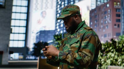 Adult male soldier in uniform navigates mobile screen on downtown street, showing disciplined confidence and respectful presence as he checks online page near urban buildings. Camera B.