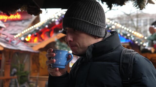 A man drinking hot cocoa at a Christmas market. Cozy winter atmosphere, festive lights and decorations in the background. Warm drink with whipped cream in hands, cold weather, holiday mood, slow