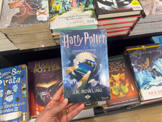 Naklejka premium BELO HORIZONTE, MG, BRAZIL, DECEMBER 12, 2025 A hand holds the iconic 'Harry Potter and the Chamber of Secrets' book in a vibrant bookstore setting, celebrating the magic of reading.