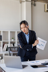 Naklejka premium Young attractive business asian woman working on laptop in the office and holding document in her hand 