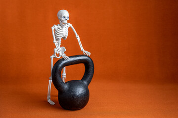 Black kettlebell on Halloween orange background with plastic skeleton, fall holiday fitness background
