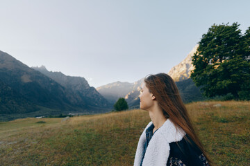 Fototapeta premium Woman in profile looks at distant mountains across a wide meadow landscape, a nature scene outdoors and travel concept with peaceful mood, long hair and layered jacket.