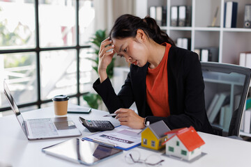 Asian business woman sitting and calculating expenses and mortgage with calculator and house on...