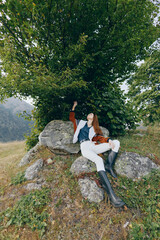 Fototapeta premium Woman relaxing on rocks under a tree in the countryside nature, wearing boots and a casual jacket, enjoying outdoor serenity on a grassy hillside near mountain scenery.