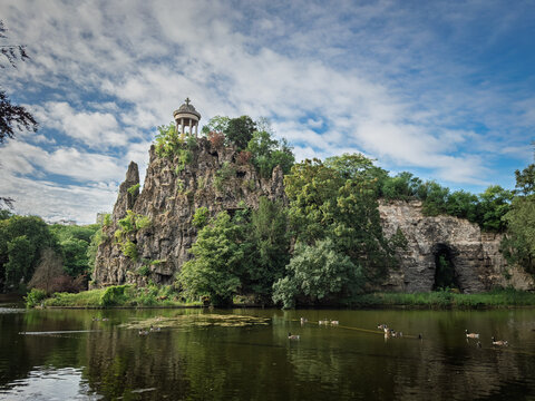 View of Ile du Belvedere in Parc des Buttes Chaumont, Paris, with the Temple de la Sybille on a rocky cliff above the lake, surrounded by lush trees and ducks swimming on the calm water.
