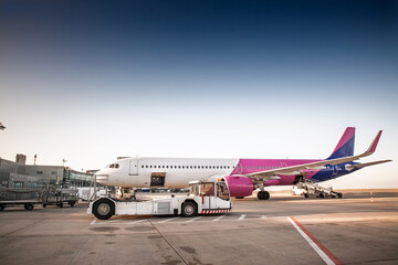 Commercial passenger airplane parked on the tarmac of a European airport, with pushback tug, service vehicles and boarding stairs positioned around the jetliner under a clear evening sky.