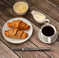 French breakfast, two fresh croissants, black coffee with cream and butter on a wooden table.