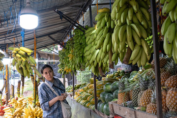 Young woman choosing fresh tropical fruits at a local fruit market stall in Sri Lanka. Pineapples, mangoes and papayas displayed on the counter with bananas hanging above.Authentic Asian marketplace.