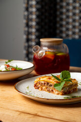 A plate of warm, cheesy lasagna sits on a rustic wooden table next to a fresh salad and a jar filled with a colorful beverage. The setting is inviting and cozy