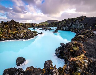 Pale blue lagoon winds amidst dark, mossy volcanic rocks under a cloudy sky