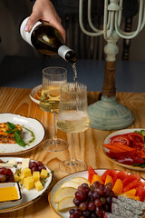 A hand pours sparkling wine into tall glasses while an array of cheeses, fresh fruits, and colorful vegetables is beautifully laid out on a rustic wooden table
