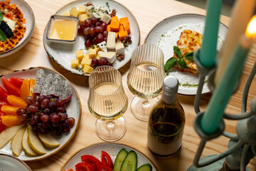 A table is beautifully arranged with various colorful fruits, artisanal cheeses, and two sparkling glasses of wine. A warm atmosphere surrounds a perfect evening meal