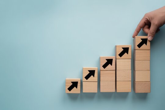 Hand stacks wooden blocks with upward arrows in a rising graph formation on a subtle blue background, symbolizing growth and success.