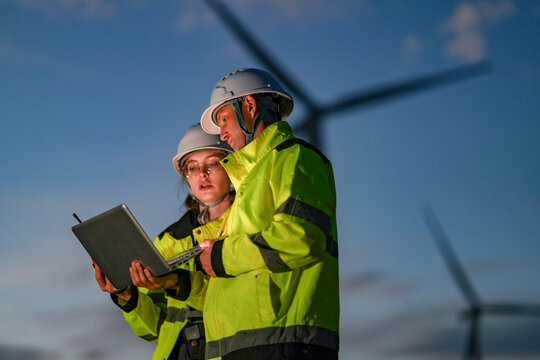 Focused female and male engineers in safety gear using a laptop at a wind farm during twilight. Technical team collaborating on a green energy project for low-carbon power.