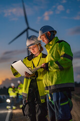 Male and female engineers in safety gear inspecting wind turbines at dusk. Female engineer holding a walkie-talkie while her colleague uses a laptop for green energy monitoring. © ultramansk