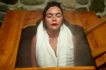 Woman with towel wrapped, eyes closed in a wooden herbal steam bath, enjoying warm