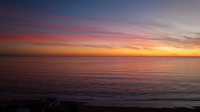 A beautiful and colorful ocean sunset as seen from Santa Cruz California looking over the pacific ocean towards a lone sailboat in a serene panning aerial drone shot with gentle waves crashing