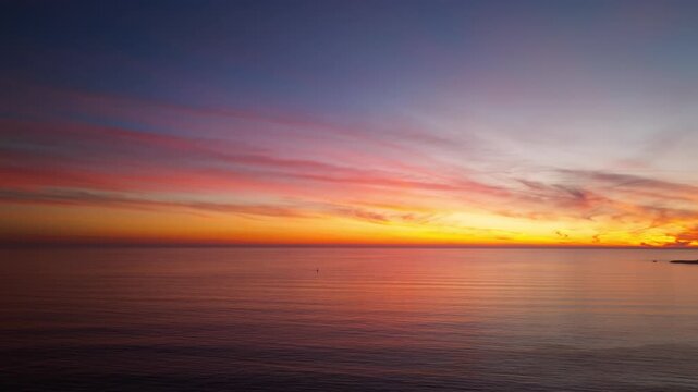 A beautiful and colorful ocean sunset as seen from Santa Cruz California looking over the pacific ocean towards a lone sailboat in a serene panning aerial drone shot with gentle waves crashing