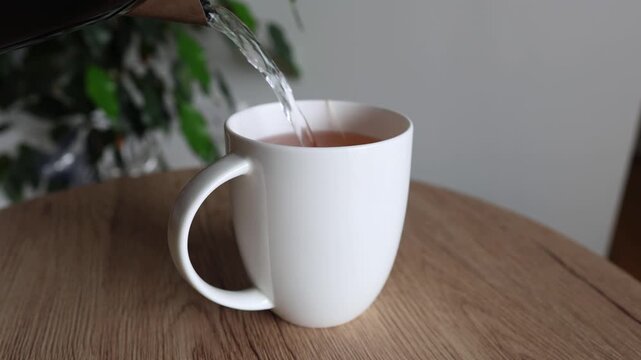 Close-up video of hot water being poured from a kettle into a white ceramic cup with a teabag on a wooden table.