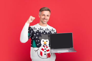 Man with laptop screen. Portrait of middle aged man in sweater isolated over red background. Concept of holidays, happiness, emotions and Christmas celebration.
