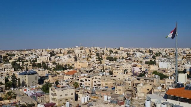 Panoramic view of Madaba, Jordan, showing rooftops with a Jordanian flag flying prominently above the city under a clear blue sky