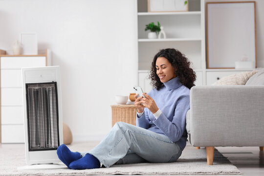 Young African-American woman with electric heater using mobile phone at home