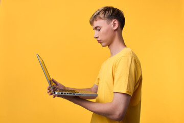 Young man in a yellow shirt studies on a laptop, standing against a vivid orange background,...