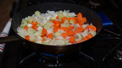 carrots, onions, and celery sizzle together in a dark skillet over the blue flame of a gas stove