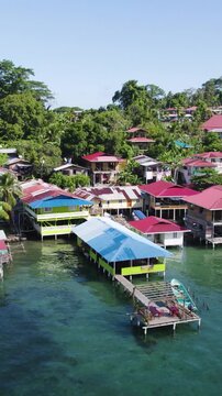 Vertical drone footage revealing a vibrant stilt house community surrounded by clear tropical waters and coastal scenery.