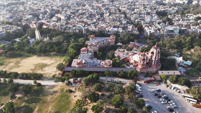 A high-angle aerial drone shot showcasing the ISKCON Temple complex in East of Kailash, Delhi. The grand red sandstone architecture of the Sri Sri Radha Parthasarathi Mandir with dense residential
