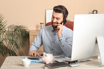 Young businessman with calculator talking by mobile phone at table in office
