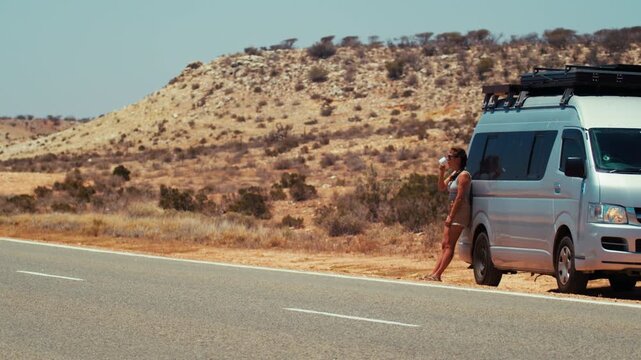 Woman traveler holds cup while leaning against van near road in arid landscape under clear sky.