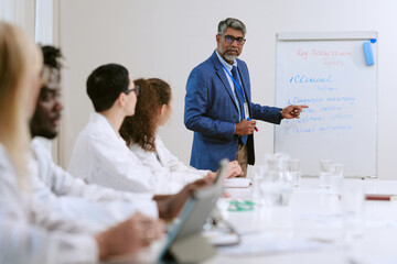 Obraz premium Middle aged Caucasian man presenting medical information on flip chart to group of young adult and middle aged multiethnic colleagues, wearing lab coats during meeting