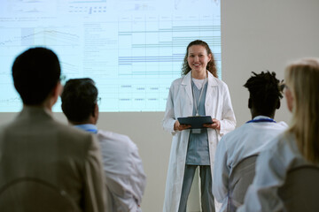 Caucasian young adult woman presenting medical data on digital tablet to diverse group of healthcare professionals during conference, standing in front of projected scientific charts
