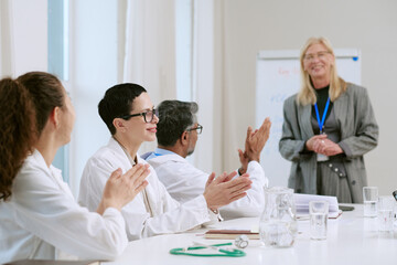 Fototapeta premium Middle aged Caucasian woman standing and smiling while presenting to diverse group of young adult and middle aged doctors, sitting at table and applauding during medical seminar