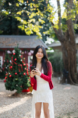 A smiling young woman wearing a Santa hat is using a phone near a Christmas tree and communicating via the internet.