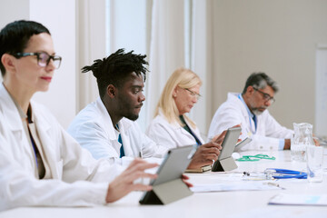 Group of diverse middle aged and senior doctors including Black man and Caucasian woman, sitting at table using digital tablets during medical conference or professional meeting