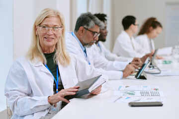Fototapeta premium Caucasian senior woman smiling while holding digital tablet, sitting at desk with diverse group of middle aged and young adult scientists working with documents and laptops