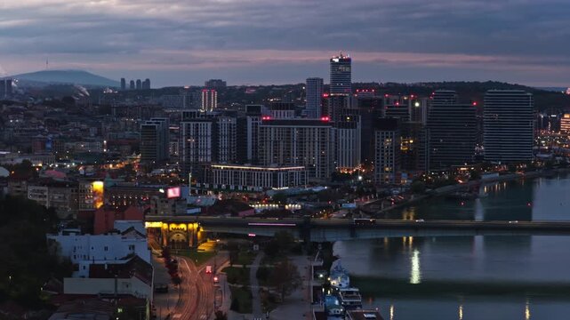 Aerial drone view of Belgrade Waterfront at blue hour, showcasing the illuminated Sava River, modern skyline, Ada Bridge, and colorful reflections on the water