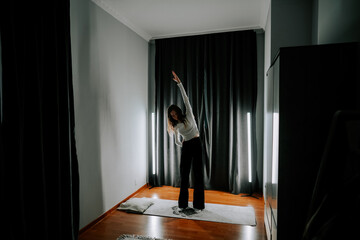 Young woman exercises at home in the evening light with curtains drawn