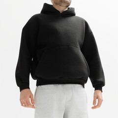 Man wears a black hoodie and gray pants while standing indoors in plain background during daytime