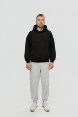 Person stands in neutral space wearing a black hoodie and gray sweatpants while looking directly at the camera