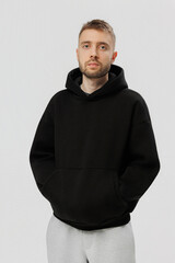 Man stands in a black hoodie and gray pants with hands in pockets against a plain background
