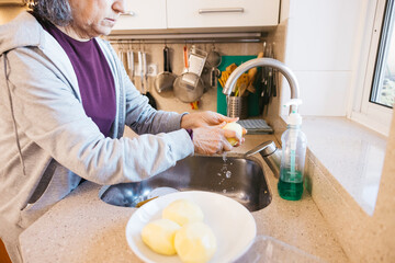 Senior woman washing fresh raw potatoes under running water in a home kitchen