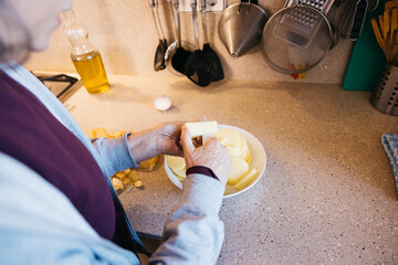 Elderly woman slicing butter, preparing potatoes for cooking on kitchen counter