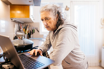 Senior woman at home kitchen preparing food while learning a new recipe online