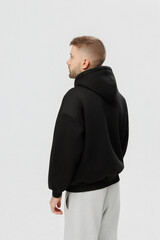 Young man stands facing away while wearing a black hoodie and gray pants in a studio setting with plain background